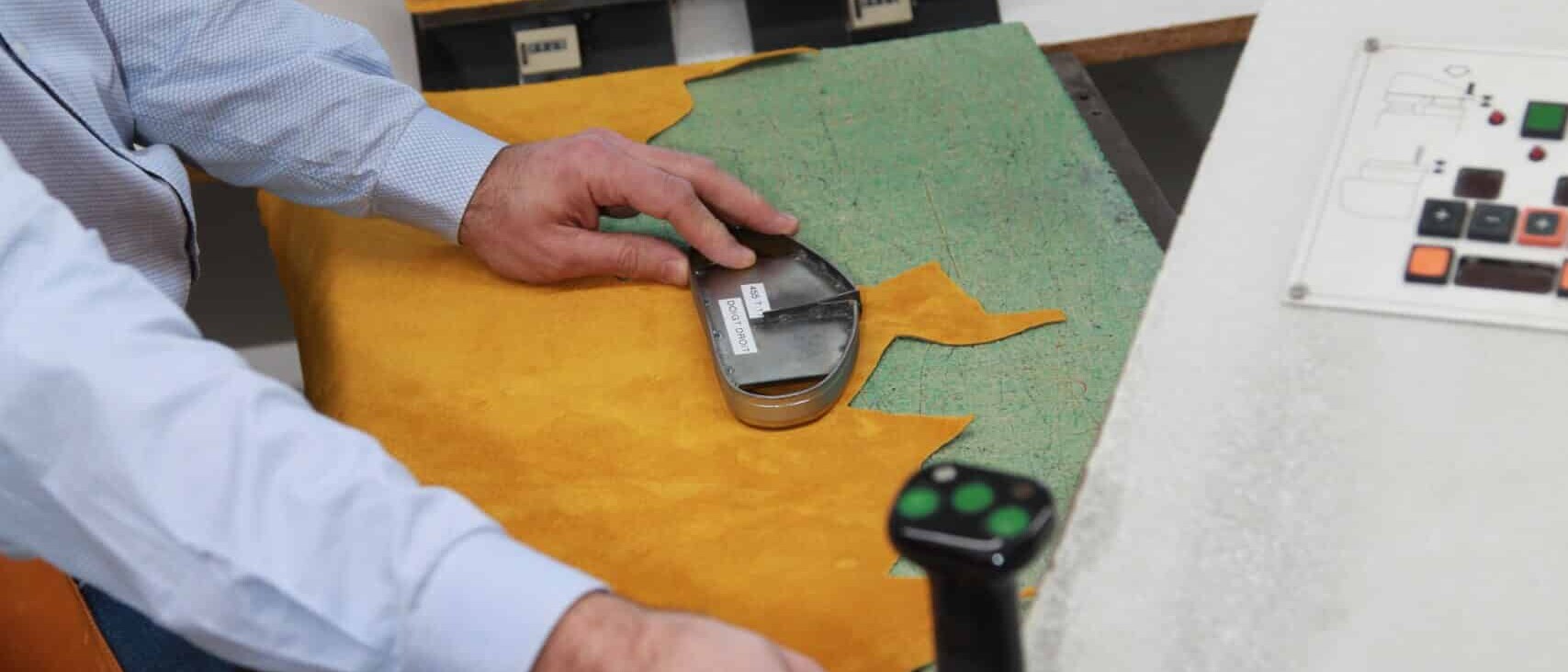 Un artisan utilise une presse industrielle pour découper du cuir à l’aide d’un emporte-pièce en métal. En arrière-plan, plusieurs matrices et morceaux de cuir préparés sont disposés sur un plan de travail.