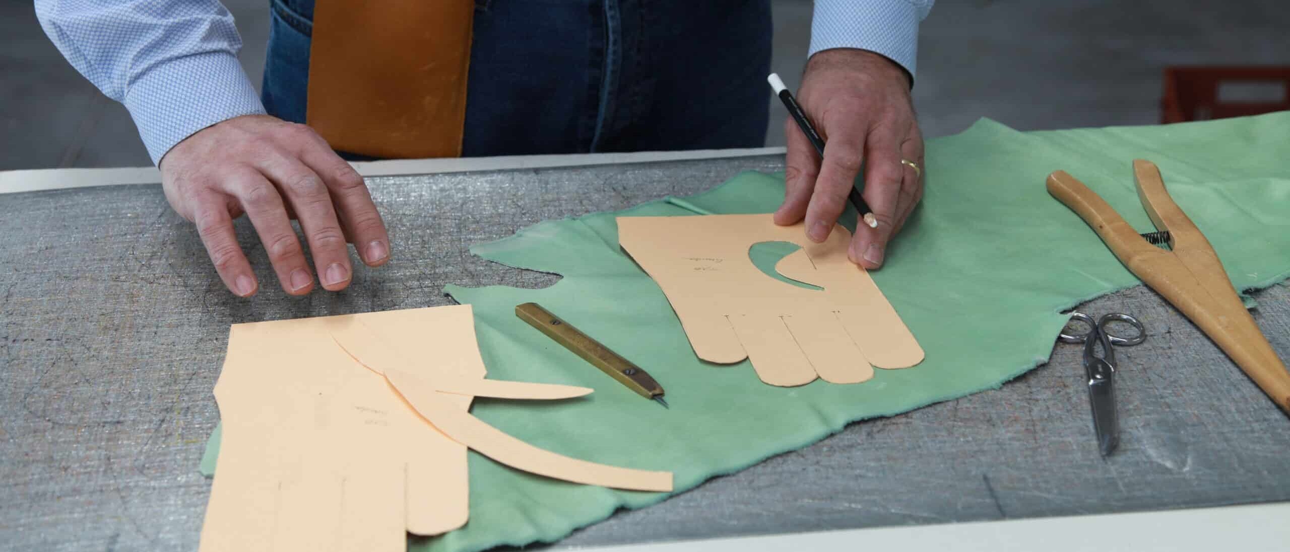 Un artisan trace le patron d’un gant sur un morceau de cuir vert à l’aide d’un gabarit en carton, avec des ciseaux, un crayon et d'autres outils de coupe posés sur la table de travail.