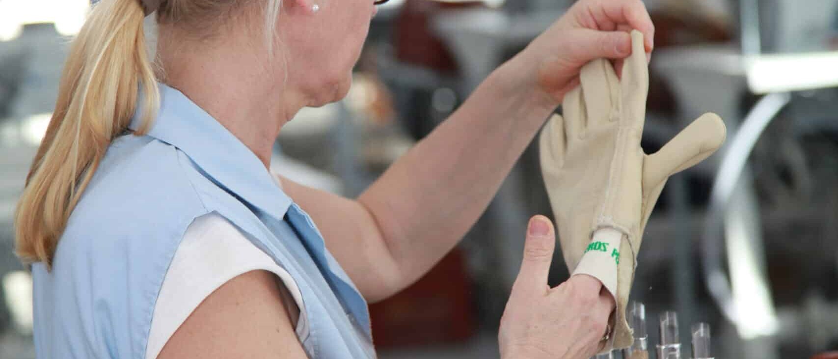 Une ouvrière en blouse bleue ajuste un gant en cuir beige sur un moule métallique dans un atelier de fabrication, vérifiant la qualité et la forme du produit.